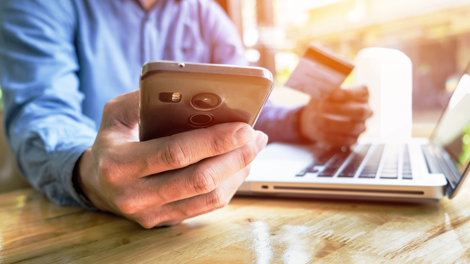 geração Y: homem em frente ao computador olhando celular e segurando cartão de crédito com a outra mão